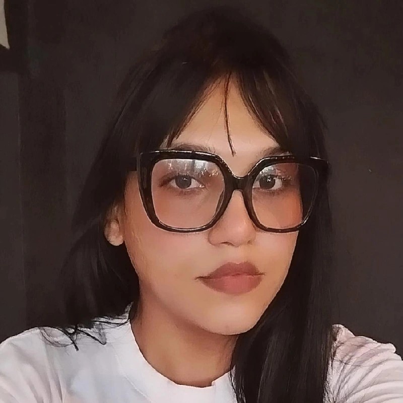 Úrsula, mulher trans de pele parda e cabelos lisos e escuros, está usando óculos com armação preta e camiseta branca.