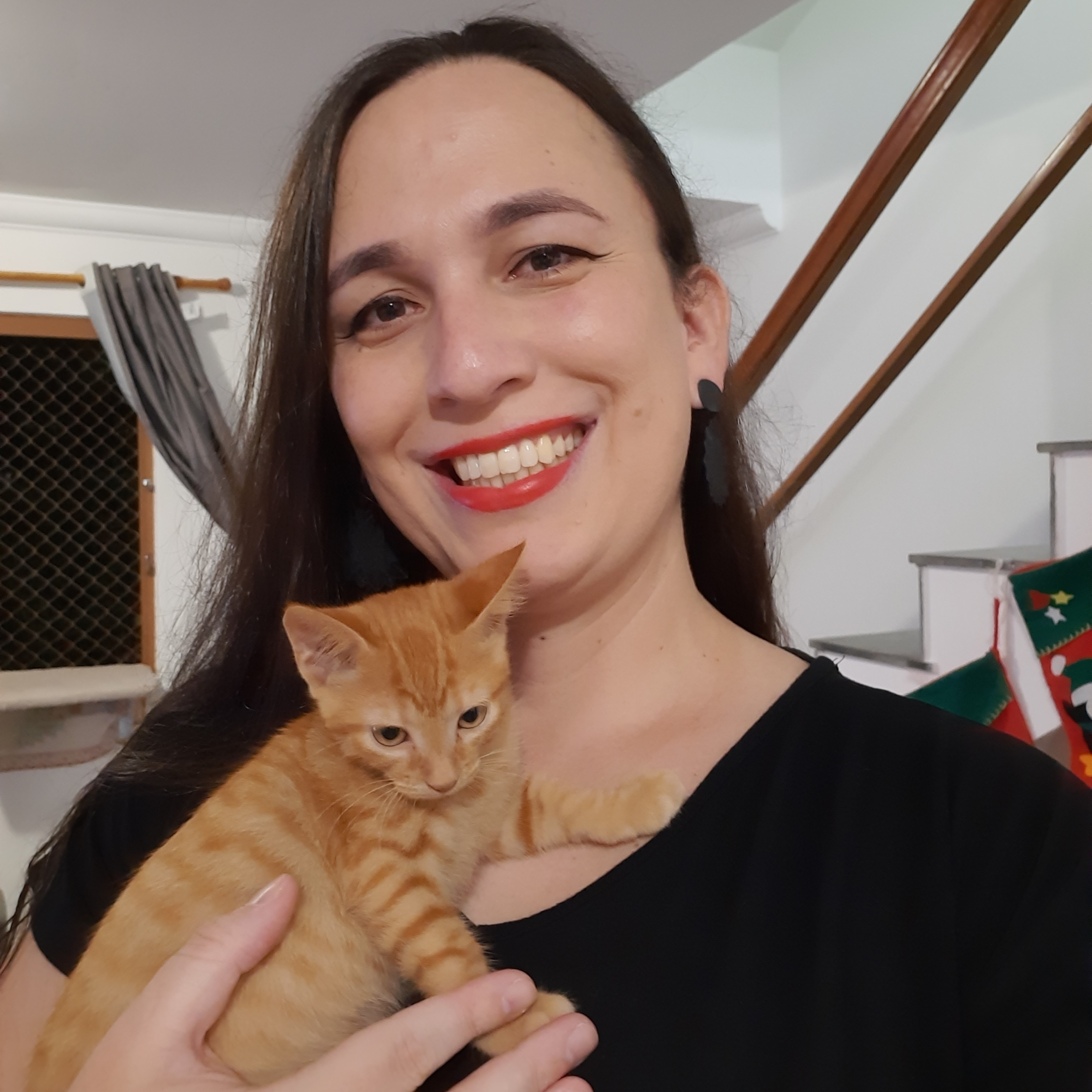 Luana, mulher branca de cabelos lisos e escuros. Veste uma camiseta preta e usa brincos pretos. Está segurando um gatinho filhote laranja.