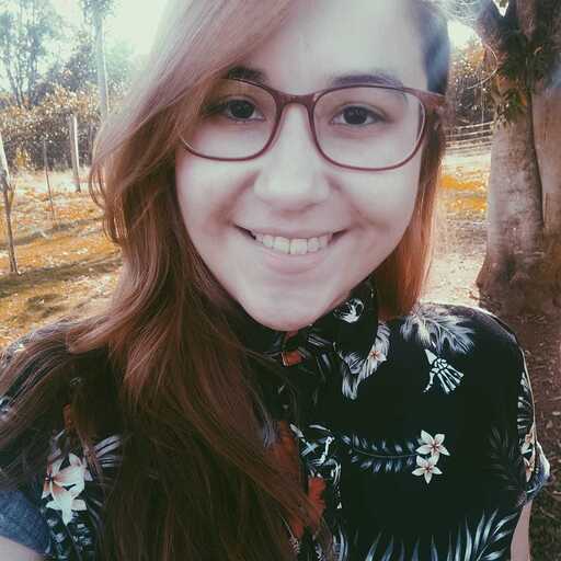 Juliana, mulher branca de cabelo liso até os ombros e ruivo. Usa óculos e possui olhos pretos, veste uma camisa com detalhes florais e tem ao fundo árvores.