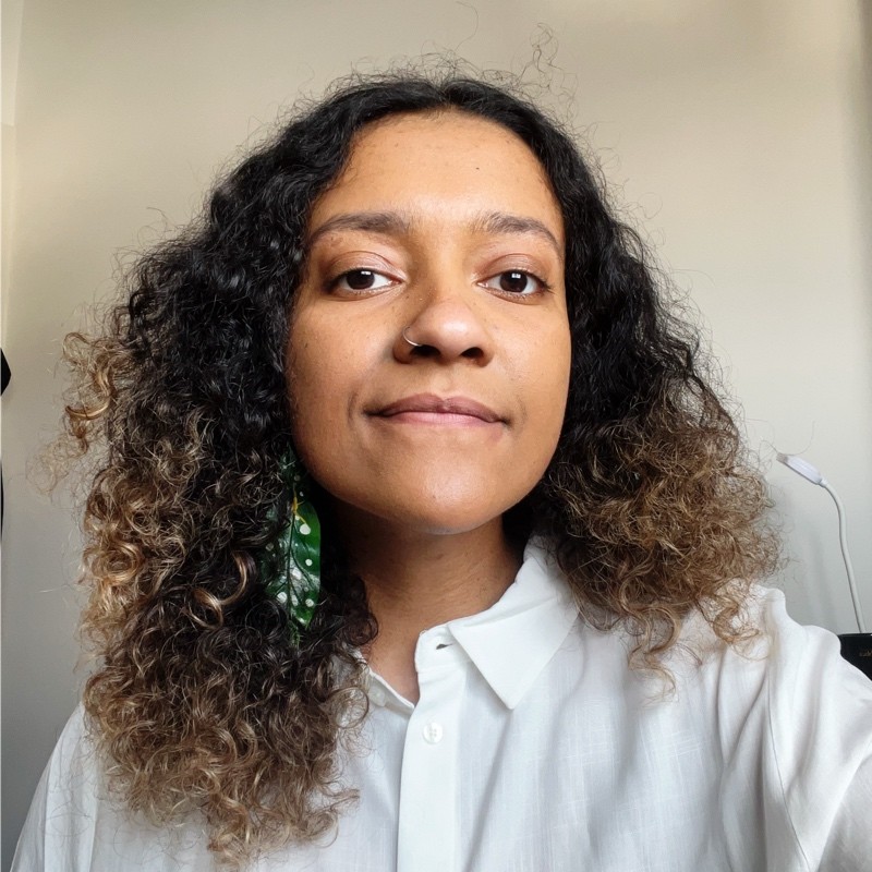 Bianca, mulher negra com cabelos cacheados, soltos e com luzes loiras na ponta. Está usando brincos verdes e camisa branca de botões.