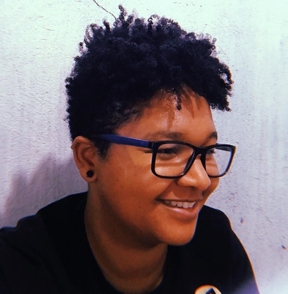 Carol, mulher negra de cabelos escuros, crespos e curto. Usa camisa preta, óculos de bordas pretas e brinco pequeno preto.