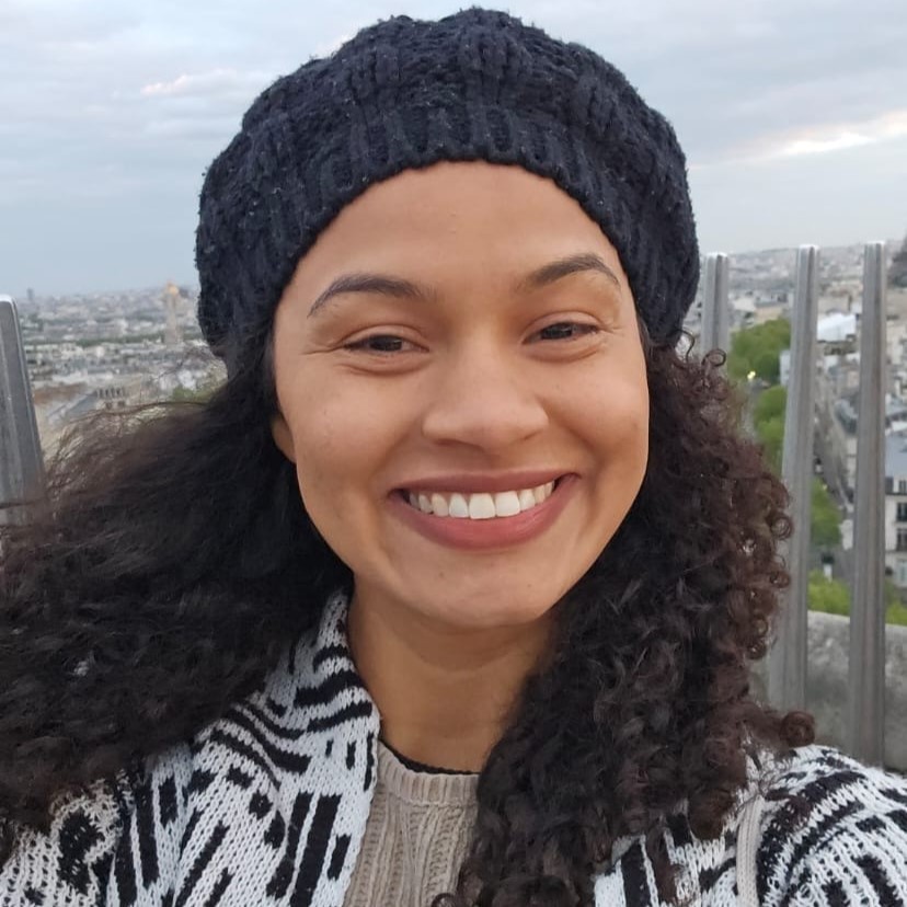 Bianca, mulher negra com cabelos cacheados, soltos e com luzes loiras na ponta. Está usando brincos verdes e camisa branca de botões.