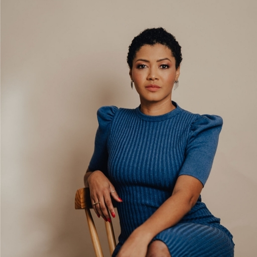 Ruth, mulher negra de cabelo curto e preto e olhos pretos amendoados. Usa conjunto azul e fundo com parede lisa bege.