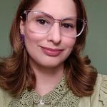 Tatiana, mulher branca de cabelos ruivos ondulados até a altura dos ombros e olhos castanhos escuros. Usa óculos com armação e lentes transparentes e camisa verde e fundo também verde.
