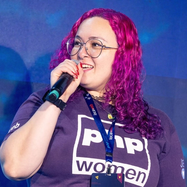 Monica, mulher branca de cabelos coloridos da cor rosa e roxo. Veste uma camiseta roxa, óculos pretos e está segurando um microfone.