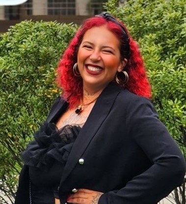 Andressa, mulher afroindígena com cabelos ondulados e vermelho vivo. Usa blusa, vestido e colar pretos, brincos de argolas grandes e douradas.