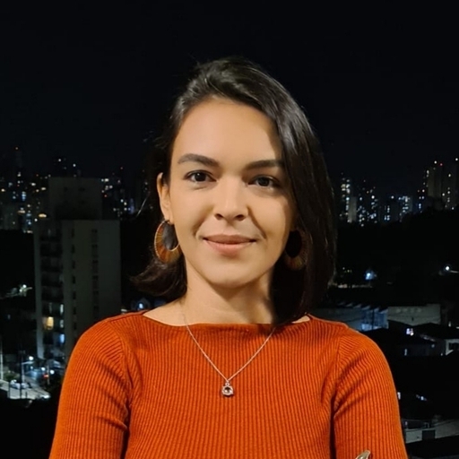 Mayra, mulher de pele parda, com cabelos lisos, escuros e na altura dos ombros. Está usando brincos de argolas marrons, camisa de cor laranja e colar prata.