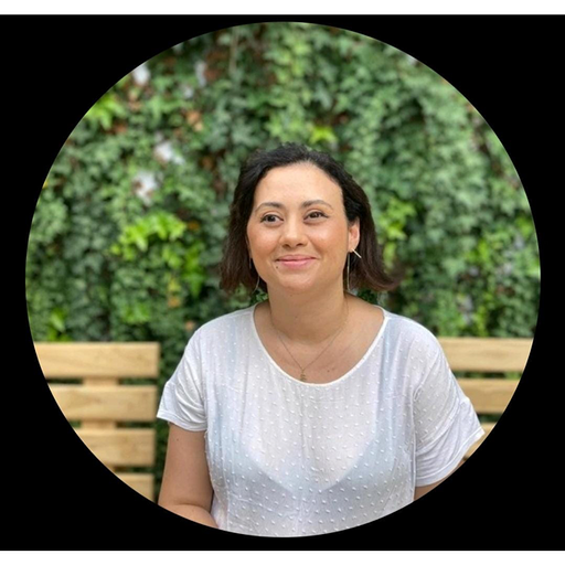 Janaína, mulher branca de cabelo ondulado ate os ombros castanhos e olhos castanho com uma camiseta branca, ao fundo de uma parede com folhas verdes.
