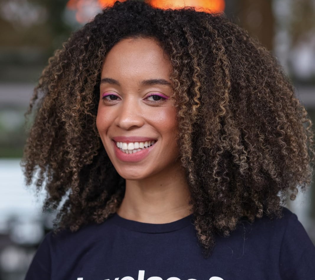 Stefani, mulher negra de cabelo cacheado com mechas castanhas. Usa uma camiseta azul marinho com os nomes das divas da programação: Lovelace, Hopper e Vaughan. O fundo é um vidro com reflexo de árvores desfocado.