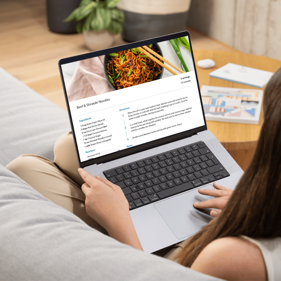 Woman sitting on a couch viewing a digital recipe for Beef & Shirataki Noodles on a laptop. The screen displays ingredients, directions, and a food photo, representing content from Whitney Morgan’s 7-Day Hashimoto’s Meal Plan.